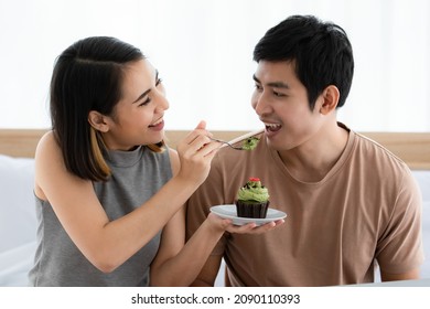 Portrait shot of cute smiling young Asian lover couple sitting on bed at home, looking at each other with romantic feeling and eating a matcha cupcake. The Wife feeds her husband with care - Powered by Shutterstock