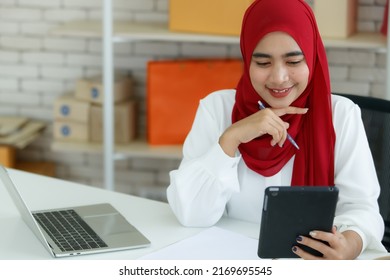 Portrait shot of cute smiling young Muslim woman wearing a red hijab sitting on a black office chair, holding digital tablet, thinking and planning about how to promote her business to clients - Powered by Shutterstock