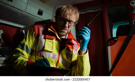 Portrait Shot Of An Emergency Care Worker Sitting In The Ambulance Car Holding A Walkie Talkie Waiting For The Call, Looking Tired And Sad, Doing A Night Shift. Paramedic At Work.	