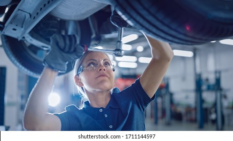 Portrait Shot of a Female Mechanic Working Under Vehicle in a Car Service. Empowering Woman Wearing Gloves and Using a Ratchet Underneath the Car. Modern Clean Workshop.