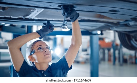 Portrait Shot Of A Female Mechanic Working On A Vehicle In A Car Service. Empowering Woman Wearing Gloves And Using A Ratchet Underneath The Car. Modern Clean Workshop.