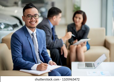 Portrait Shot Of Handsome Asian Car Dealer Wearing Elegant Suit Looking At Camera With Wide Smile While Distracted From Working Meeting At Showroom