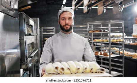 Portrait shot of handsome Caucasian male in white apron and hat standing in front of camera, holding tray with sweet bakery and smiling in kitchen of bakehouse. Man worker of bakeshop. - Shutterstock ID 2251396711