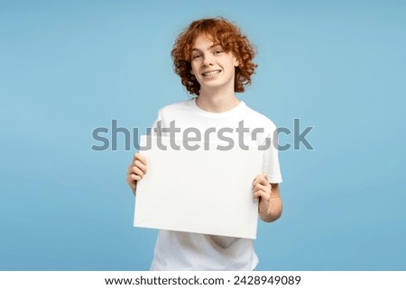 Portrait shot of a handsome curly haired red head male teen, smiling with braces and holding a banner, set against a blue studio background. Mockup concept