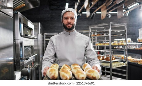 Portrait shot of handsome male in white apron and hat standing in front of camera, holding tray with fresh just-baked bagueattes and smiling in kitchen of bakehouse. Man in bakeshop showing bread. - Shutterstock ID 2251396691