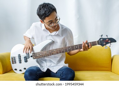 Portrait shot of handsome smiling young teenager enjoying playing the bass guitar. Junior guitarist in casual clothes sitting on the yellow couch and holding an instrument while looking at camera - Powered by Shutterstock