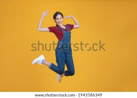 Portrait shot of happy beautiful Asian woman in denim dungarees. She jumping by proud in herself isolated on yellow background.