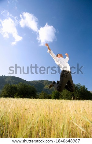 A portrait shot of a happy businessman jumping in the air with his arms outstretched in the field.