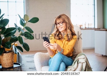 Portrait shot of happy mature woman using her mobile phone and text messaging while relaxing at home in the armchair. 