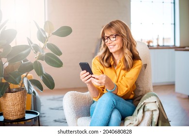 Portrait Shot Of Happy Middle Aged Woman Using Mobile Phone And Text Messaging While Relaxing In The Armchair At Home.