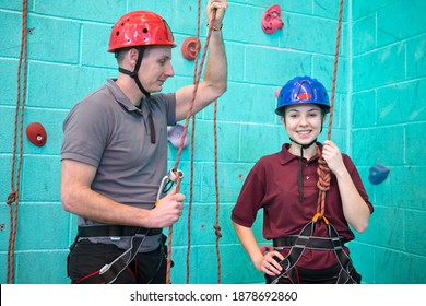 Portrait Shot Of A High School Student In Harness Standing With A Gym Teacher Against A Rock Climbing Wall.