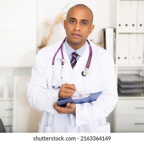 Portrait Shot Of Latin American Male Doctor Holding Clipboard In Hand While Standing In Office