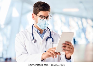 Portrait shot of male doctor wearing face mask and using digital tablet while standing in hospital’s foyer.  - Powered by Shutterstock