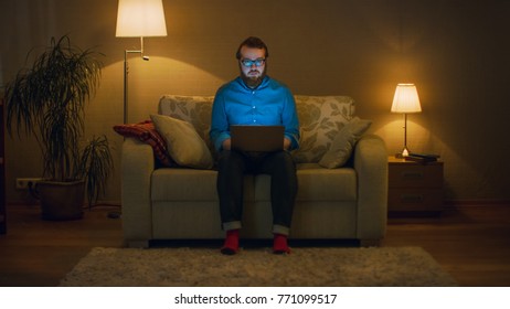 Portrait Shot Of A Man Sitting On A Couch In His Living Room, Laptop On His Knees. He's Working. He Looks Straight At The Camera. Floor Lamps Are Turned On.