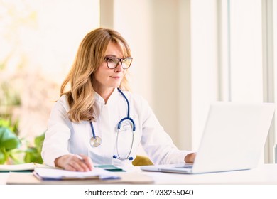 Portrait shot of middle aged female doctor sitting at desk and working on laptop in doctor’s office. - Powered by Shutterstock