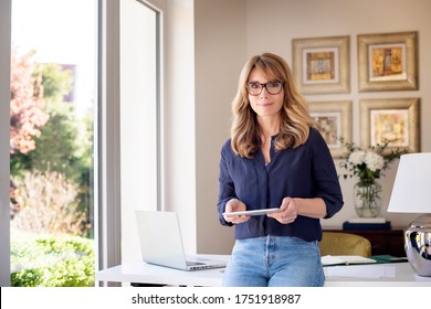 Portrait Shot Of Middle Aged Woman Holding Digital Tablet In Her Hand While Looking At Camera And Smiling. Smiling Businesswoman Working From Home. Home Office.