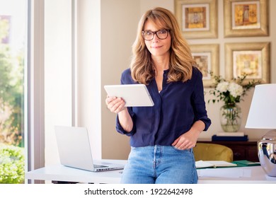 Portrait Shot Of Middle Aged Woman Holding Digital Tablet In Her Hand While Looking At Camera And Smiling. Smiling Businesswoman Working From Home. Home Office.