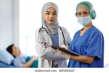 Portrait Shot Of Muslim Islam Arab Doctor In Hijab White Lab Coat With Stethoscope And Asian Professional Female Nurse In Blue Hospital Uniform With Face Mask And Goggles Standing Smiling Together.