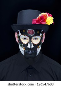 Portrait Shot Of A Scary Man Wearing Traditional Sugar Skull Make-up And Hat Decorated With Roses Over Plain Black Background.