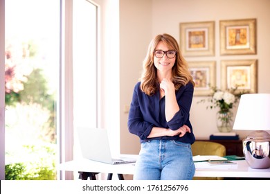 Portrait Shot Of Smiling Mature Woman Standing At Desk At Home While Looking At Camera And Smiling. 