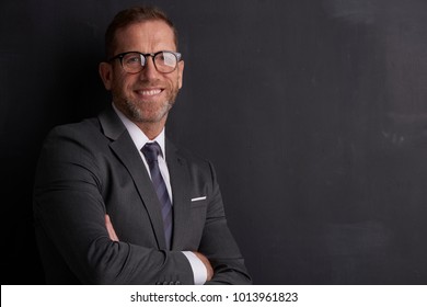 Portrait Shot Of A Smiling Middle Aged Man Wearing Suit And Standing With Arms Crossed At Dark Background. 