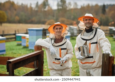 Portrait shot of smiling middle aged female beekeeper in protective suit and adult male colleague looking at camera while standing at entrance to apiary farm next to autumn forest, copy space - Powered by Shutterstock