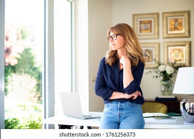 Portrait shot of thinking mature woman standing at desk at home while looking out the window.  - Powered by Shutterstock