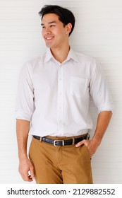 Portrait shot of young adult Asian confident man with cute smiling wearing a formal white long sleeve shirt and brown pants with hand in pocket posturing on background in studio - Shutterstock ID 2129882552