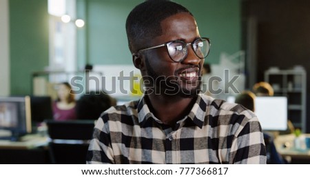 Portrait shot of young African American man in glasses turning his head to the camera and smiling in the modern office. Working people on the blurred background. Close up