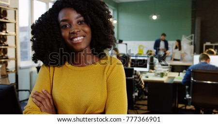 Portrait shot of young attractive curly African American woman smiling into the camera in the modern office. Working people on the blurred background. Close up.