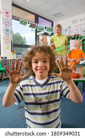 A Portrait Shot Of A Young Smiling Boy Showing His Dirty Muddy Hands To The Camera With Teacher And Other Students Standing In Background.