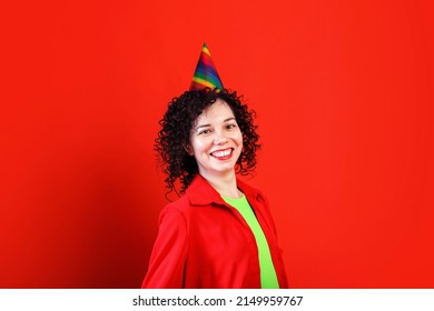 Portrait Of Shy Curly Haired Girl In Red Shirt And Colorful Party Hat. Positive Young Female, Looking Cheerfully In Camera And Smiling Widely. Indoor Studio Shot Isolated On Red Background.