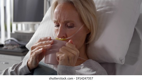 Portrait Of Sick Aged Woman Patient With Breathing Tube Drinking Water In Mug. Senior Ill Female Resting In Bed At Hospital Ward And Holding Cup With Water Or Tea