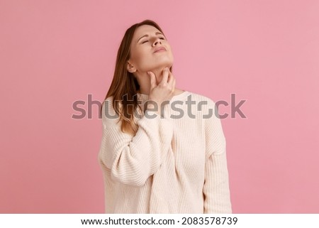 Portrait of sick blond woman touching sore neck feeling unwell, suffering tonsils inflammation, medical concept, wearing white sweater. Indoor studio shot isolated on pink background.