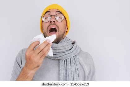 Portrait of sick handsome man wearing grey sweater, yellow hat and spectacles, blowing nose and sneeze into tissue. Male have flu, virus or allergy respiratory. Healthy, medicine and people concept - Powered by Shutterstock