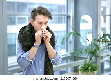 Portrait Of Sick Ill Handsome Guy In Scarf Blowing His Nose In Paper Handkerchief Indoors At Office Building, Young Man With Runny Nose, Flu. Symptoms Of Coronavirus, Covid-19. Allergy, Feeling Unwell