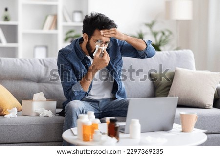 Portrait Of Sick Indian Guy Blowing Nose In Paper Tissue At Home, Ill Young Eastern Man Feeling Fever, Suffering Seasonal Flu Or Cold, Having Rhinitis Or Allergic Reaction, Closeup Shot
