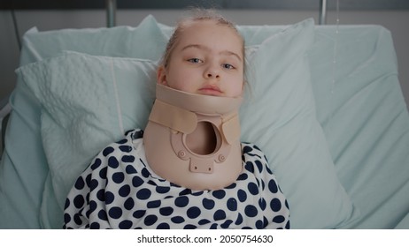 Portrait Of Sick Little Patient Resting In Bed Looking At Camera While Having Neck Cervical Collar Recovering After Painful Surgery In Hospital Ward. Child Wearing Oxygen Nasal Tube During Examination