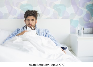 Portrait Of Sick Man With Thermometer In Mouth Reclining On Bed At Home