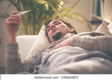 Portrait of sick men lying on a sofa and putting his hand on his head, holding thermometer. Young man has high fever, takes his temperature and coughs. Epidemic of influenza and coronavirus, covid-19