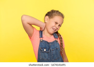 Portrait Of Sick Unhappy Little Girl With Braid In Denim Overalls Massaging Painful Back, Feeling Discomfort In Neck, Child Sedentary Lifestyle Concept. Studio Shot Isolated On Yellow Background