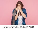 Portrait of sick unhealthy brown haired woman suffering runny nose flu symptoms virus grippe, wearing checkered shirt and scarf. Indoor studio shot isolated on pink background.