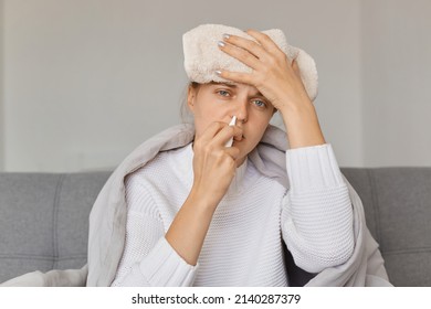 Portrait Of Sick Upset Woman With Wet Towel On Her Forehead Being Wrapped In Blanket Sitting On Sofa And Using Nasal Spray, Treating Runny Nose, Having High Temperature.