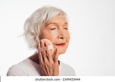 Portrait With Side View Of Old Woman With Grey Hair Doing Skincare And Tretmant With Sponge. Take Care About Good Looking. Rubbing Skin On Face. Make Up. Isolated Over White Background