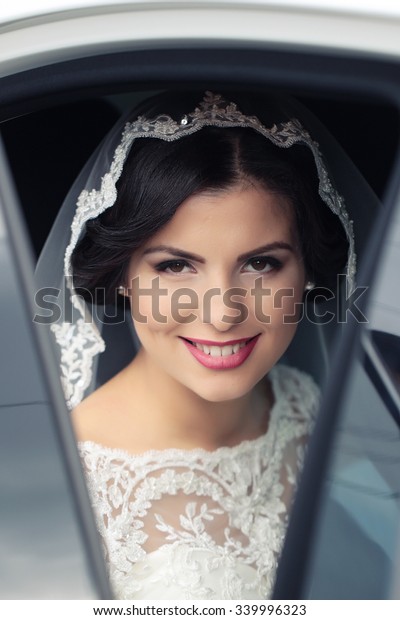 Portrait with side view of one\
beautiful young brunette sensual smiling bride in white lace dress\
and veil on head looking forward sitting in car, vertical\
picture