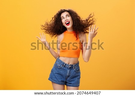 Portrait of silly positive and energized cute female with curly hairstyle in cropped top flicking hair strand and smiling charming gazing at upper right corner dreamy posing happy over orange wall