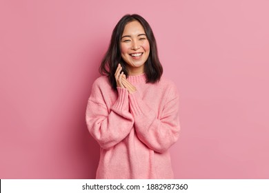 Portrait of sincere brunette Asian female model rubs palms and smiles broadly expresses happiness feels joyful has white even teeth wears oversized casual sweater isolated over pink background