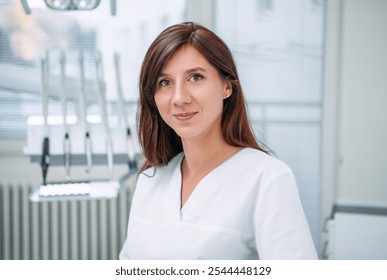 Portrait of sincerely smiling young dentist woman dressed white medical scrubs uniform sitting in modern dental clinic next to stomatology chair. Health care and medicare industry concept image. - Powered by Shutterstock