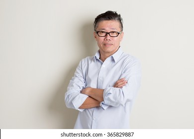 Portrait of single mature 50s Asian man in casual business arms crossed smiling and standing over plain background with shadow.