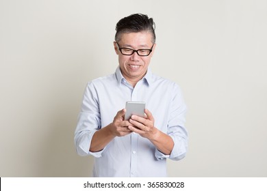 Portrait Of Single Mature 50s Asian Man In Casual Business Using Mobile Apps On Smartphone And Smiling, Standing Over Plain Background With Shadow. Chinese Senior Male People.